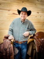 A man wearing a black cowboy hat and plaid shirt stands in front of a wooden wall, holding onto two saddles.